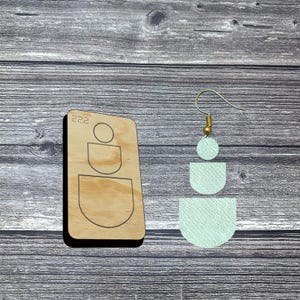 May include: A wooden die-cut with the number "222" and a geometric design, next to a mint green earring with a gold-colored hook. The earring has a circular top, a half-circle, and a larger half-circle shape.