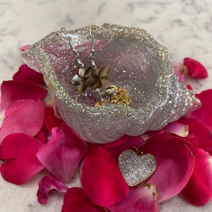 May include: A silver glitter resin dish holds a pair of flower earrings and a gold necklace. A gold and silver heart pendant rests on a bed of bright pink rose petals. The dish is surrounded by more petals.