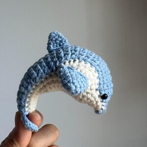 Könnte beinhalten: Ein handgehäkelter Delfin in Hellblau und Weiß. Der Delfin hat einen gebogenen Körper, eine Rückenflosse und ein kleines schwarzes Perlenauge. Die Handarbeit zeigt die Textur des Garns.