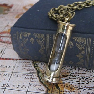 May include: A gold-toned hourglass pendant necklace with a clear glass hourglass. The necklace is on a dark blue book with gold accents.