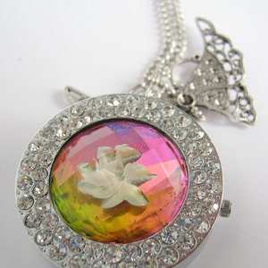 Puede incluir: Un collar de plata con un colgante redondo que presenta una flor blanca dentro de una cúpula de vidrio iridiscente multicolor rodeada de diamantes de imitación transparentes.