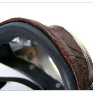 May include: Close-up of a brown leather saddle with a black trim. The saddle has a curved shape and a textured surface. The image is set against a white background.