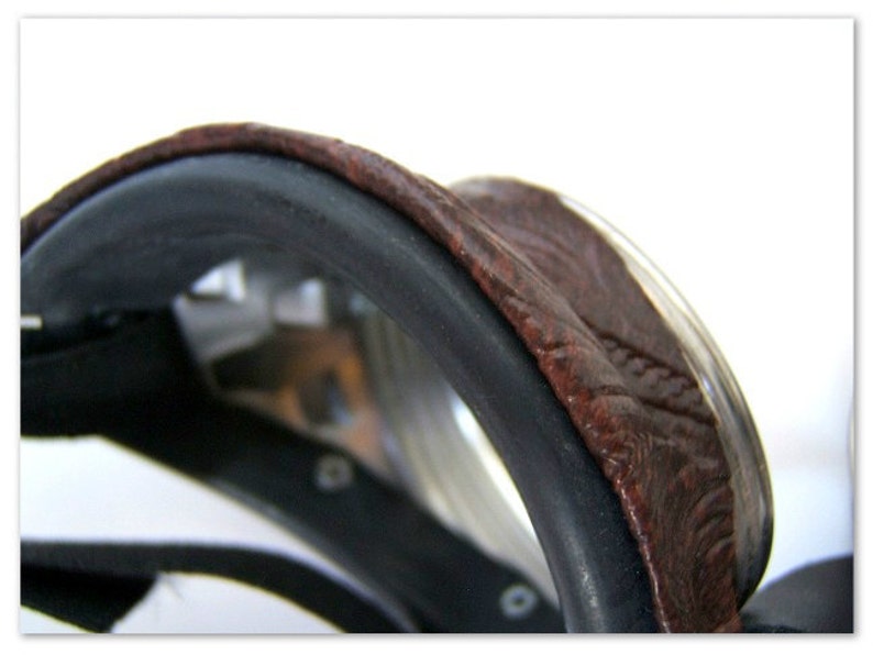 May include: Close-up of a brown leather saddle with a black trim. The saddle has a curved shape and a textured surface. The image is set against a white background.