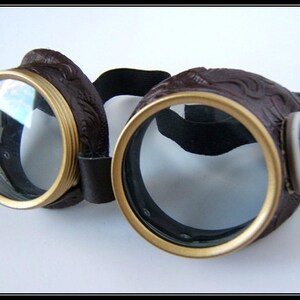 May include: Brown steampunk goggles with gold-colored rims and clear lenses. The goggles have a decorative embossed design on the leather frames and a black elastic strap. A cameo is on the side of the goggles.
