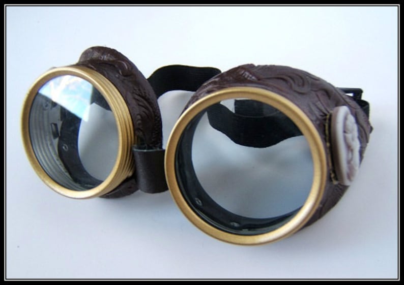 May include: Brown steampunk goggles with gold-colored rims and clear lenses. The goggles have a decorative embossed design on the leather frames and a black elastic strap. A cameo is on the side of the goggles.