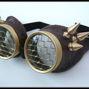 May include: Brown leather steampunk goggles with gold-colored metal accents and a mesh lens. The goggles have multiple gold-colored spikes on the side.