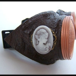 May include: Brown steampunk goggles with copper-colored rims and a decorative cameo. The goggles have a textured, embossed design and a black adjustable strap. The lenses have a grid pattern.