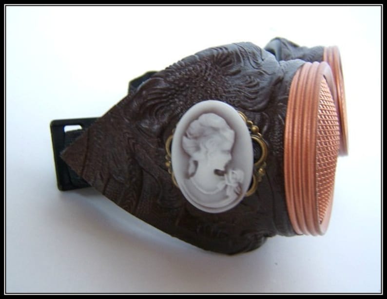 May include: Brown steampunk goggles with copper-colored rims and a decorative cameo. The goggles have a textured, embossed design and a black adjustable strap. The lenses have a grid pattern.