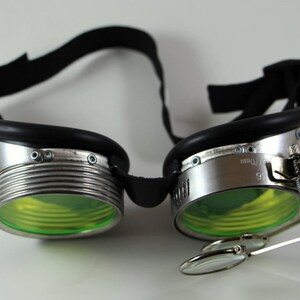 May include: Silver steampunk goggles with green lenses and a magnifying glass attached to the side. The goggles have a black leather strap.