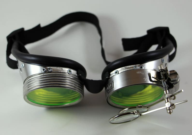 May include: Silver steampunk goggles with green lenses and a magnifying glass attached to the side. The goggles have a black leather strap.