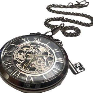 Puede incluir: Un reloj de bolsillo plateado con esfera esqueleto, mostrando los intrincados mecanismos internos. El reloj tiene números romanos y una cadena a juego. Un dije decorativo en forma de llave cuelga, agregando un toque vintage.