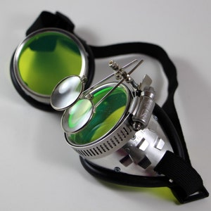 May include: Silver steampunk goggles with green tinted lenses and a black adjustable strap. The goggles have two smaller magnifying lenses attached to the side.