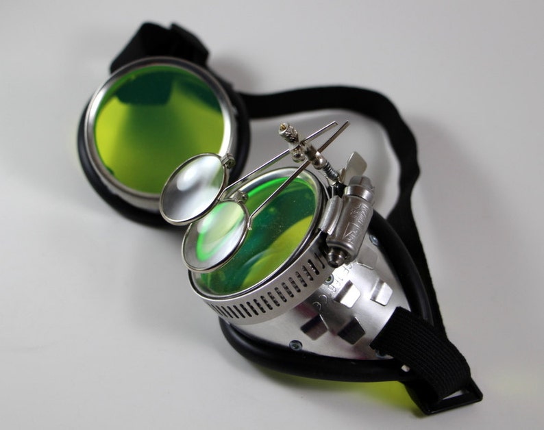 May include: Silver steampunk goggles with green tinted lenses and a black adjustable strap. The goggles have two smaller magnifying lenses attached to the side.