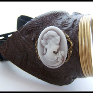 May include: Brown steampunk goggles with a decorative cameo and gold-colored metal accents. The goggles feature a dark brown leather-like material with an embossed pattern and a black adjustable strap. The lenses are surrounded by gold-colored metal rings.