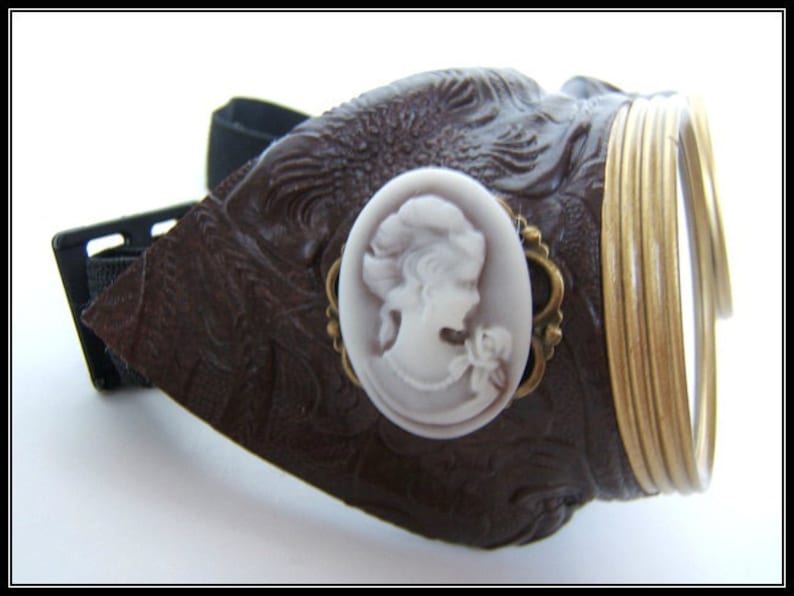 May include: Brown steampunk goggles with a decorative cameo and gold-colored metal accents. The goggles feature a dark brown leather-like material with an embossed pattern and a black adjustable strap. The lenses are surrounded by gold-colored metal rings.