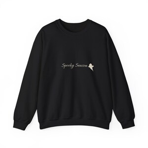 Könnte beinhalten: Ein schwarzes Langarm-Sweatshirt mit Rundhalsausschnitt, das auf der Brustmitte den Schriftzug Spooky Season in weißer Schreibschrift sowie eine kleine weiße Geistergrafik rechts neben dem Text aufweist.