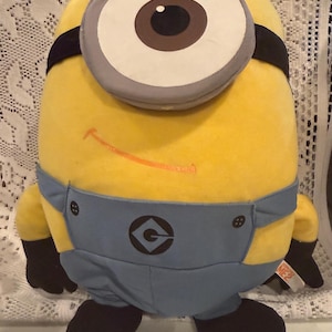 以下が含まれることがあります： 大きな目が一つ付いたミニオンのキャラクターの形をした黄色いぬいぐるみ。灰色のゴーグルのフレーム、黒いストラップ、細いオレンジ色の笑顔が特徴です。黒いロゴが入った青いデニム風のオーバーオールを着ており、手と足は黒色です。