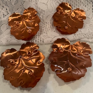 May include: A set of four metallic copper-colored dishes shaped like leaves. Each dish features a detailed embossed vein pattern and a scalloped edge, with a reflective, polished finish that highlights the curves and contours of the leaf design.