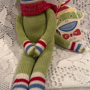 May include: A green knit sock monkey doll wearing a red scarf and a yellow and red hat. The monkey has a white face with a blue mouth and striped hands and feet in red, white, and blue. A tag attached to the monkey features a graphic of the monkey face and the text Jo Jo.