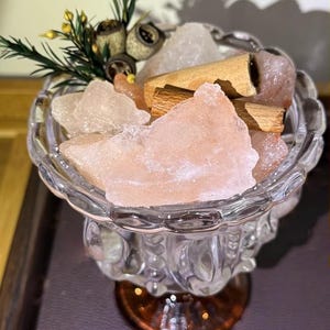Puede incluir: Un cuenco de vidrio transparente lleno de cristales de sal rosas y blancos y palitos de canela. El cuenco está adornado con ramitas de vegetación y descansa sobre una base marrón. El texto "Mature Au Boutique" es visible.