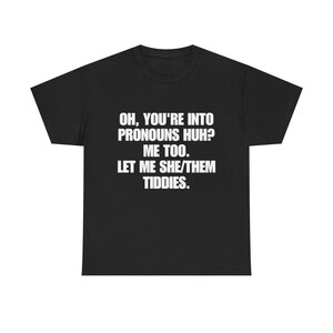 Pode incluir: Uma t-shirt preta de manga curta com texto branco impresso na frente que diz: OH, YOU'RE INTO PRONOUNS HUH? ME TOO. LET ME SHE/THEM TIDDIES. A peça tem um decote redondo e um corte padrão sobre um fundo preto sólido.