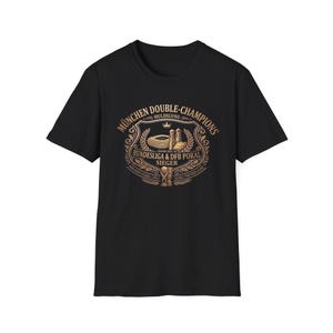 May include: Black t-shirt featuring a gold emblem with the text "München Double-Champions" and "Bundesliga & DFB Pokal Sieger". The emblem includes illustrations of a stadium, towers, and a trophy. The shirt is a short-sleeved crew neck.