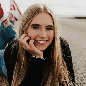 May include: A young woman with long blonde hair is lying on her stomach and smiling at the camera. She is wearing a black long-sleeved shirt and blue jeans. Her red sneakers are visible in the background. The text "Charlotte Oliver" is written in red cursive at the top of the image.