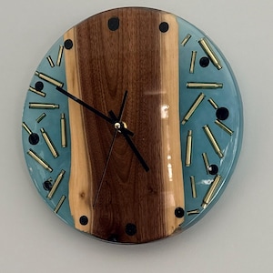 May include: A circular wall clock featuring a central vertical slab of dark brown wood with natural live edges. The surrounding area is filled with translucent light blue resin embedded with scattered brass bullet casings and small black circular markers.
