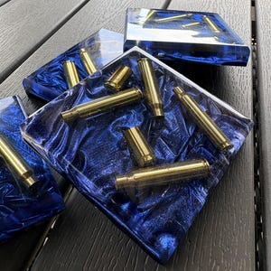 May include: A set of square coasters made of clear resin with a deep blue, crinkled, metallic-effect background. Each coaster is embedded with multiple brass-colored metal bullet casings of varying lengths arranged in a scattered pattern.
