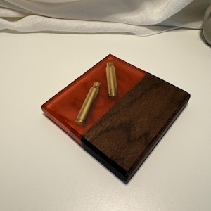 May include: A square coaster featuring a combination of dark brown wood and translucent orange resin. Two brass-colored metal bullet casings are embedded within the resin section, creating a distinct contrast against the smooth wood grain.