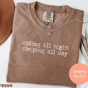 May include: A brown short-sleeved t-shirt with a crew neckline featuring the white text "coding all night sleeping all day" printed on the front. The shirt has a Comfort Colors brand label on the inside collar indicating a size small.