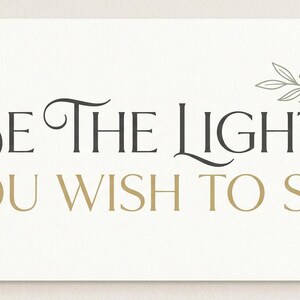 May include: A rectangular card with the text "BE THE LIGHT YOU WISH TO SEE" in black and gold lettering. A decorative floral element is in the upper right corner. The background is a light cream color.