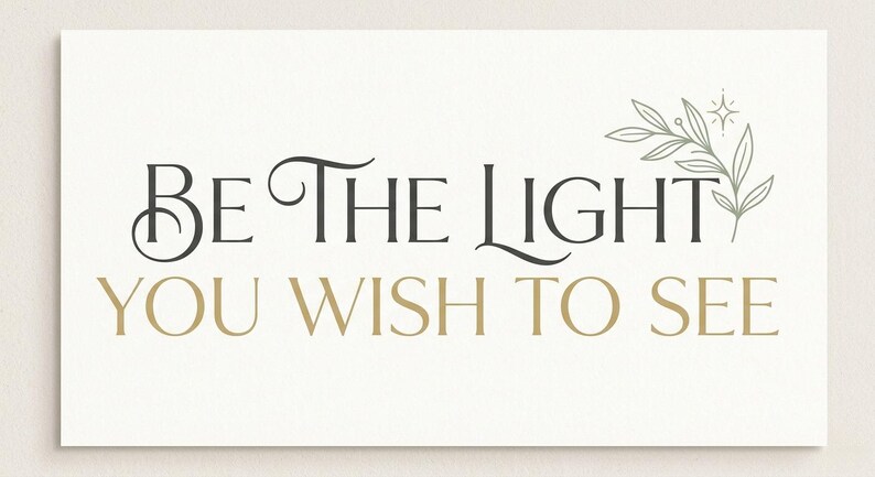 May include: A rectangular card with the text "BE THE LIGHT YOU WISH TO SEE" in black and gold lettering. A decorative floral element is in the upper right corner. The background is a light cream color.