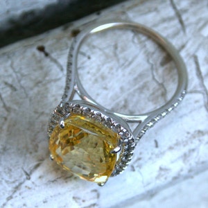 Op de afbeelding: Een zilveren ring met een grote, kussenvormige gele edelsteen. De edelsteen is omgeven door een halo van kleinere, heldere stenen. De ringband is ook versierd met kleine, heldere stenen. De ring ligt op een getextureerd, wit oppervlak.