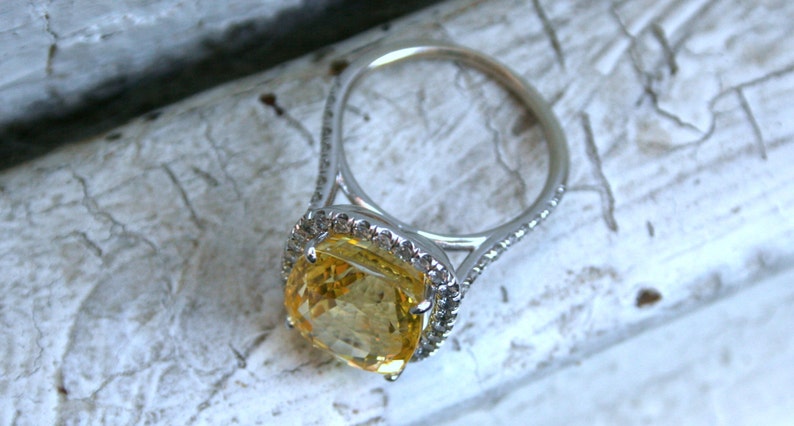 Op de afbeelding: Een zilveren ring met een grote, kussenvormige gele edelsteen. De edelsteen is omgeven door een halo van kleinere, heldere stenen. De ringband is ook versierd met kleine, heldere stenen. De ring ligt op een getextureerd, wit oppervlak.