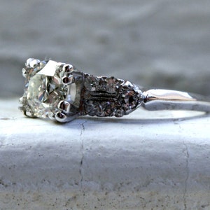 May include: A silver engagement ring featuring a large, clear, round diamond set in a prong setting. The band is adorned with smaller diamonds.