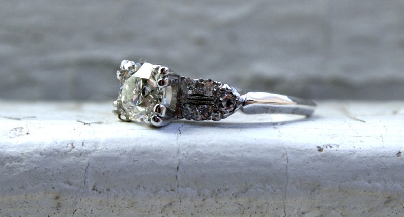 May include: A silver engagement ring featuring a large, clear, round diamond set in a prong setting. The band is adorned with smaller diamonds.