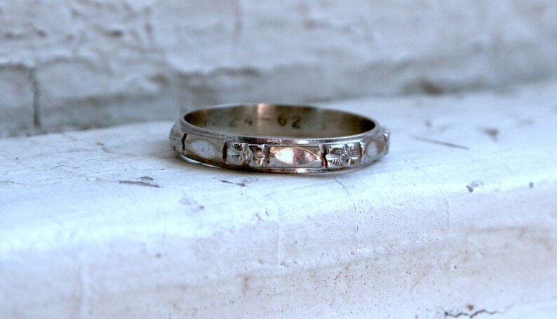 May include: A silver wedding band with a decorative pattern of small flowers and ovals. The band is inscribed with the date "24-62".
