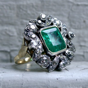 May include: A vintage emerald and diamond ring featuring a large, green emerald centre stone surrounded by a halo of smaller diamonds. The ring is set in silver and has a gold band.