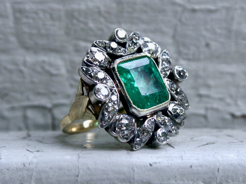 May include: A vintage emerald and diamond ring featuring a large, green emerald centre stone surrounded by a halo of smaller diamonds. The ring is set in silver and has a gold band.