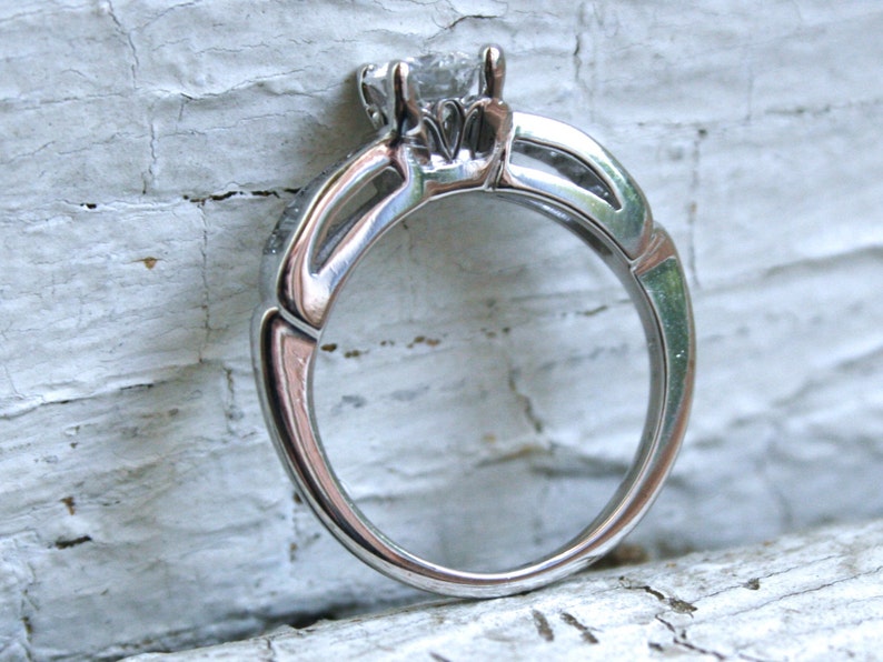 K&ouml;nnte beinhalten: Ein silberner Verlobungsring mit einem einzelnen, klaren, runden Brillanten in einer Krappenfassung. Die Ringschiene ist in einem zarten, gedrehten Design gehalten.