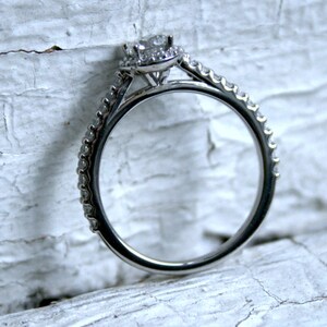 May include: A silver engagement ring with a single round diamond surrounded by smaller diamonds. The ring has a thin band with small diamonds set along the edge.