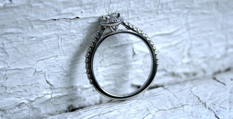 May include: A silver engagement ring with a single round diamond surrounded by smaller diamonds. The ring has a thin band with small diamonds set along the edge.