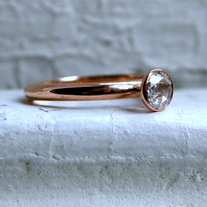May include: Rose gold ring with a clear, round gemstone. The ring has a smooth, polished band and a bezel setting for the gemstone. The ring is displayed on a textured, white surface.