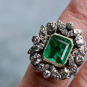 May include: A vintage ring featuring a large emerald green gemstone surrounded by a halo of smaller diamonds. The ring is set in silver and gold.