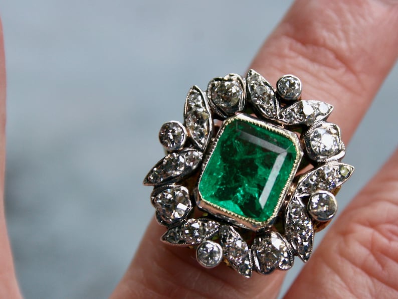May include: A vintage ring featuring a large emerald green gemstone surrounded by a halo of smaller diamonds. The ring is set in silver and gold.