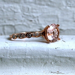 May include: A rose gold engagement ring with a large oval-shaped pink gemstone surrounded by smaller diamonds. The band is adorned with a delicate row of smaller diamonds.
