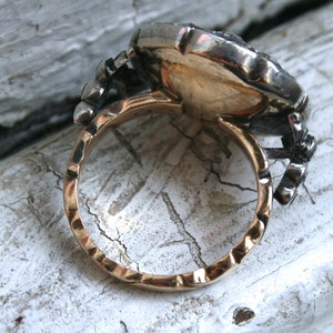 May include: A gold and silver ring with a large, round, faceted stone set in the center. The ring has a textured band and a decorative design on the side.