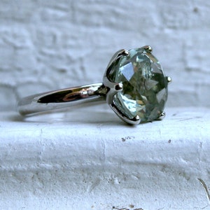 May include: A silver ring featuring a large, oval-cut, pale green gemstone. The gemstone is set in a prong setting, and the band is smooth and polished. The ring is displayed against a white, textured background.