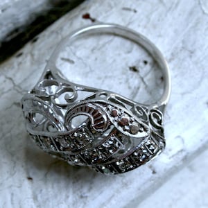 May include: A silver ring with a delicate, intricate design featuring a filigree pattern and small, clear stones. The ring is set against a white, textured background.